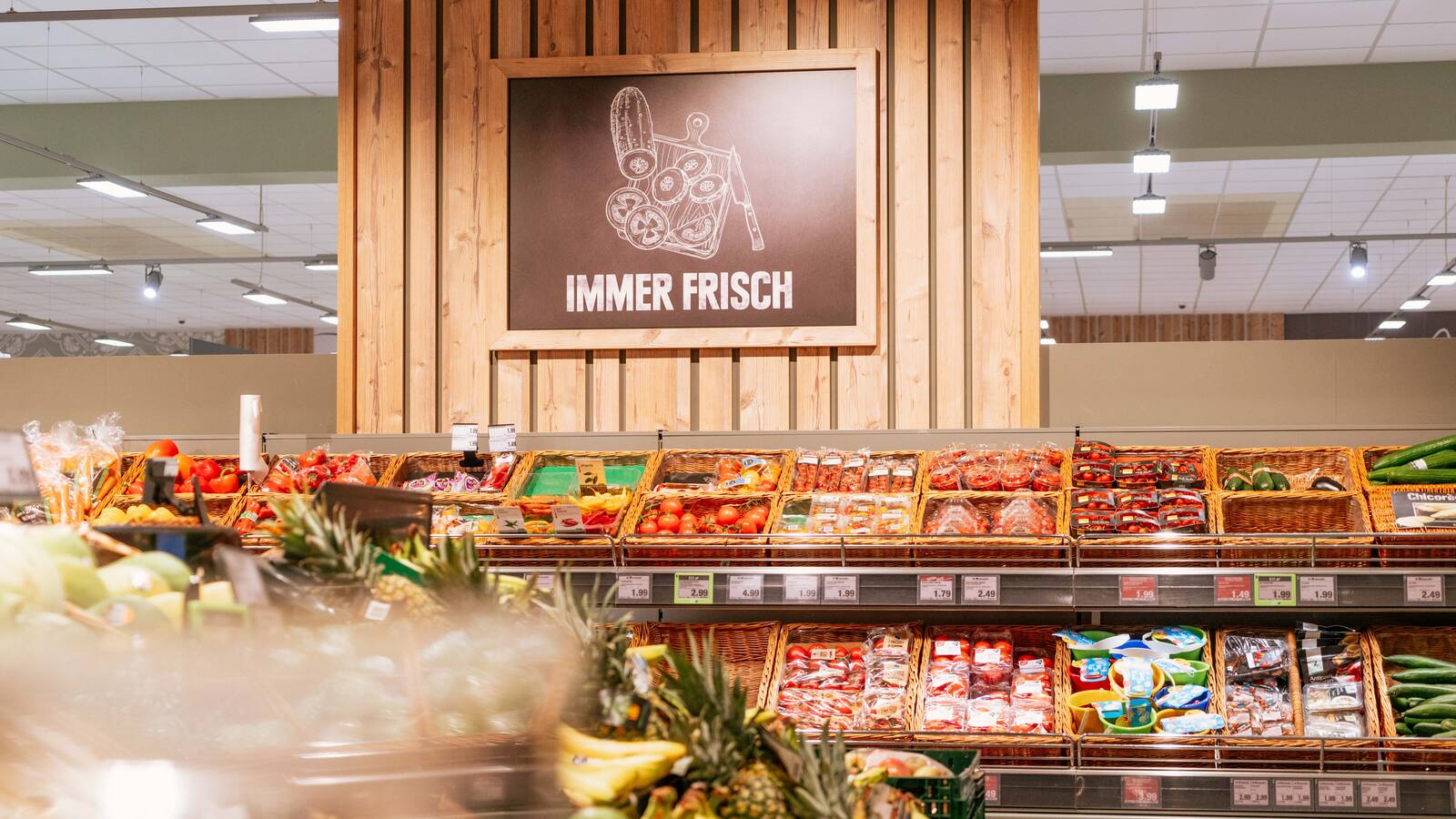 Über dem Gemüseregal des Supermarktes sind Holzpaneel mit dem Bild einer geschnittenen Gurke und Text: Immer frisch.