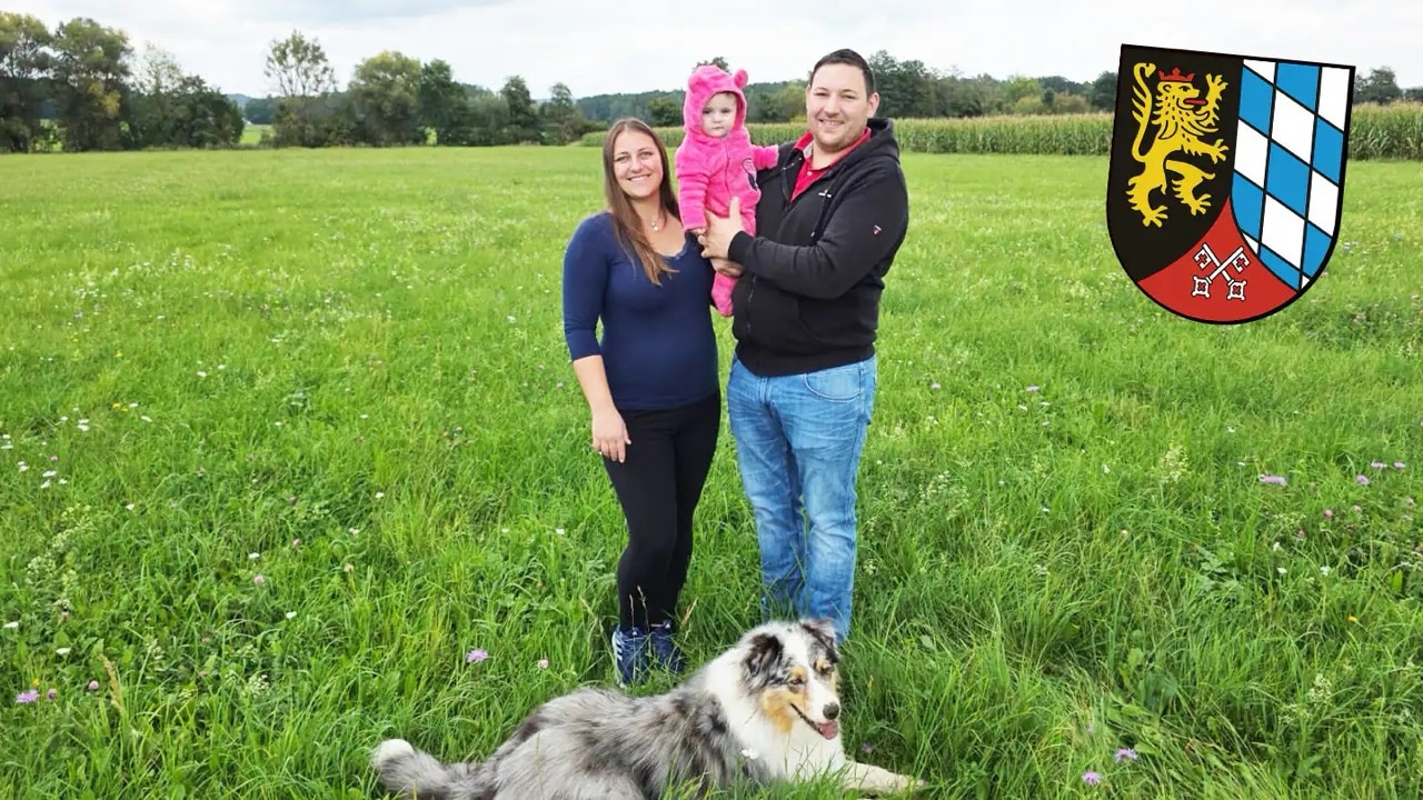 Ein Mann und eine Frau stehen mit einem Säugling auf einem Feld. Vor ihnen liegt ein Hund.