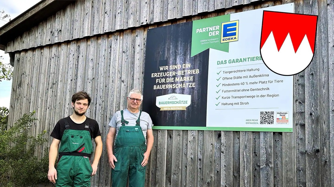 Ein jüngerer und ein älterer Mann stehen vor einer Scheune, an der ein großes Schild befestigt ist: Wir sind ein Erzeuger-Betrieb für die Marke Bauernschätze.