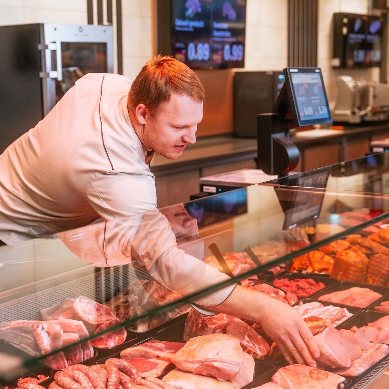 Ein Mann in Metzgerkleidung greift in die Fleisch-Auslage hinter dem Tresen.