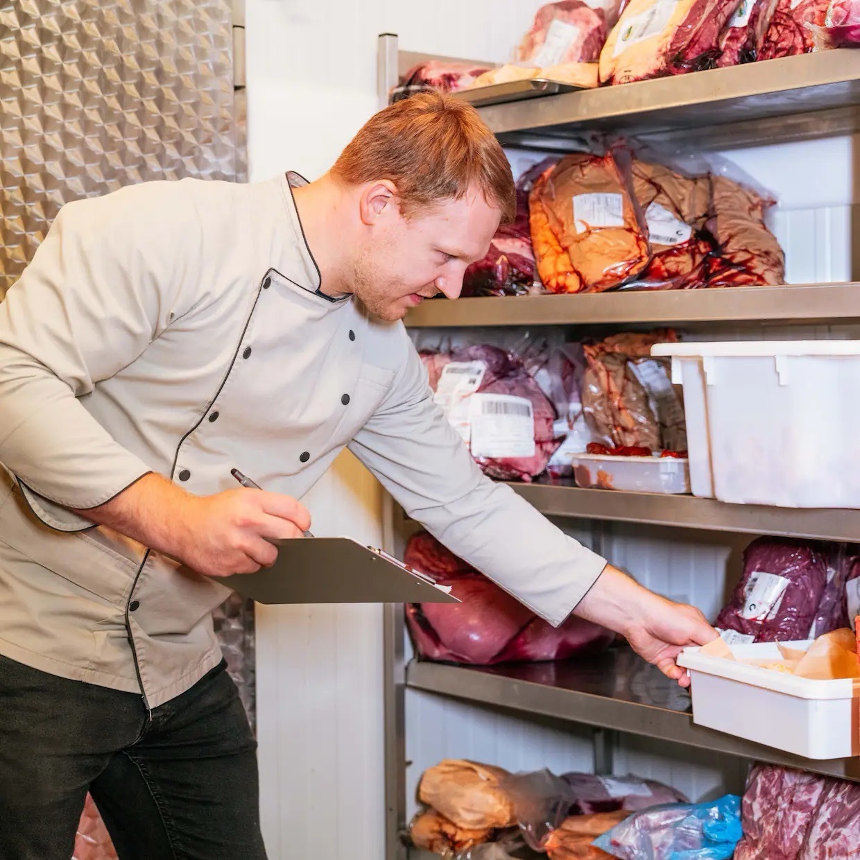 Ein Mann in Metzgerkleidung steht an einem Regal und hält ein Schreibbrett sowie einen Stift.