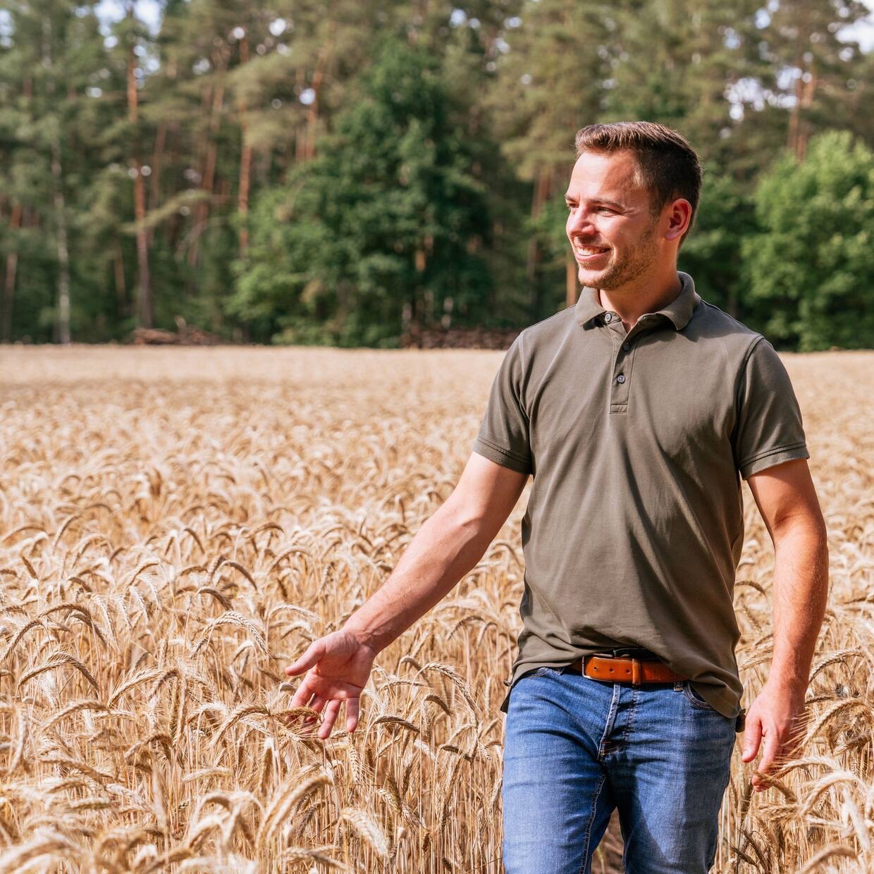 Ein Mann in braunem Poloshirt geht lächelnd durch ein Weizenfeld.