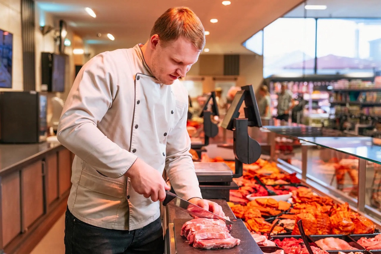 Ein Mann in Metzgerkleidung schneidet hinter dem Tresen ein Stück Fleisch in Scheiben.