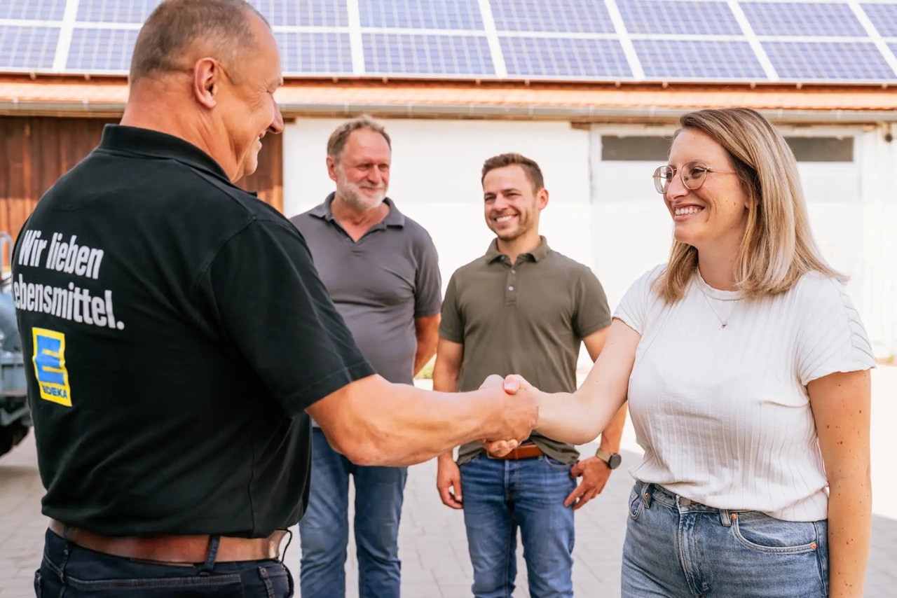 Eine Frau mit Brille gibt einem Herrn in schwarzem EDEKA-Poloshirt die Hand und lacht.