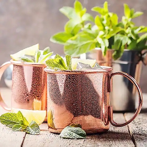 Zwei glänzende kupferfarbene Tassen, aus denen Zitronenzesten und grüne Blätter herausragen.