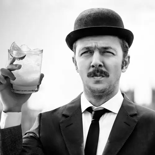 Schwarz-weiß-Bild eines Herrn mit einem Bowlerhut, einem schwarzen Anzug mit weißem Hemd und einem Drink in der erhobenen Hand.
