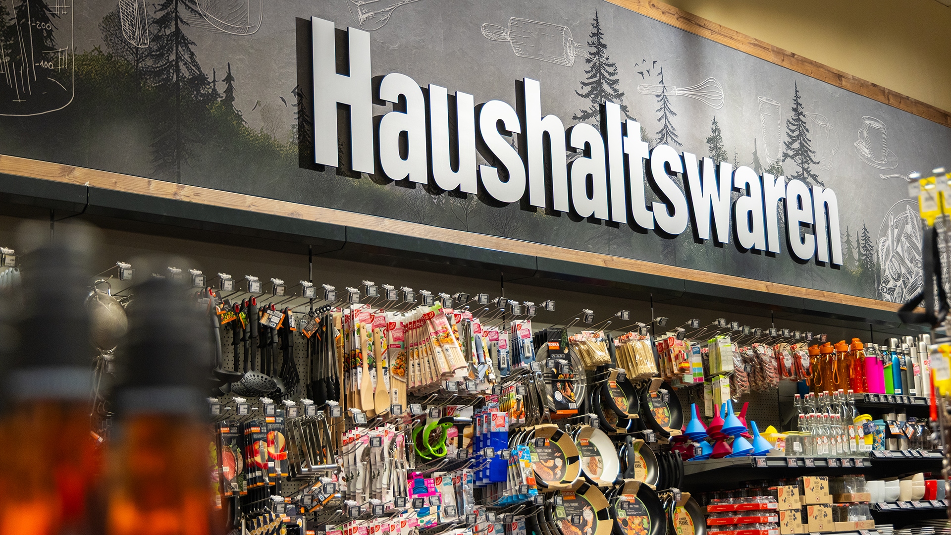 Eine Wand mit verschiedenen Haushaltswaren der Marke EDEKA zuhause und GUT&Günstig. Darüber ein großes Wandschild "Haushaltswaren".