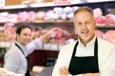 Zwei Männer mit schwarzen Schürzen stehen hinter einer Fleischtheke.