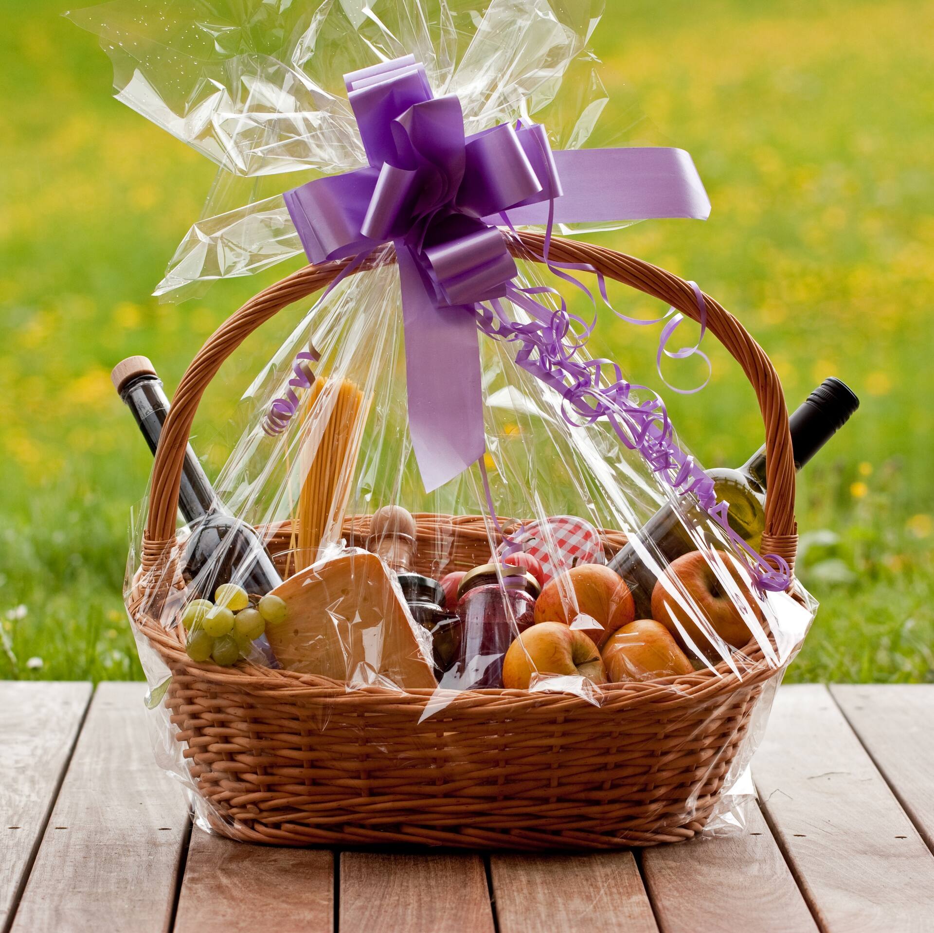 Präsentkorb mit lilaner Schleife. Unter der Klarsichtfolie liegen Äpfel, Marmelade, Käse, Weintrauben, Öl und eine Flasche Wein.