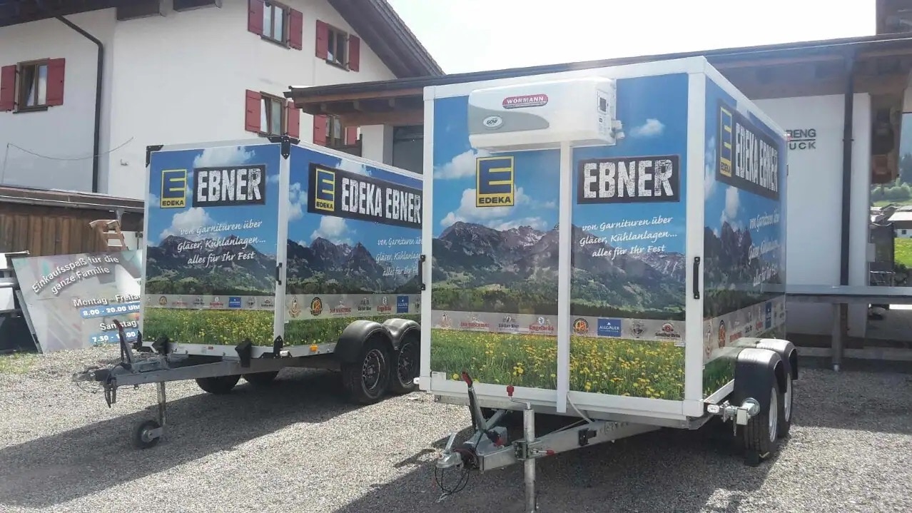 Zwei bunt gestaltete Anhänger mit einer Berglandschaft sowie EDEKA-EBNER-Logo.
