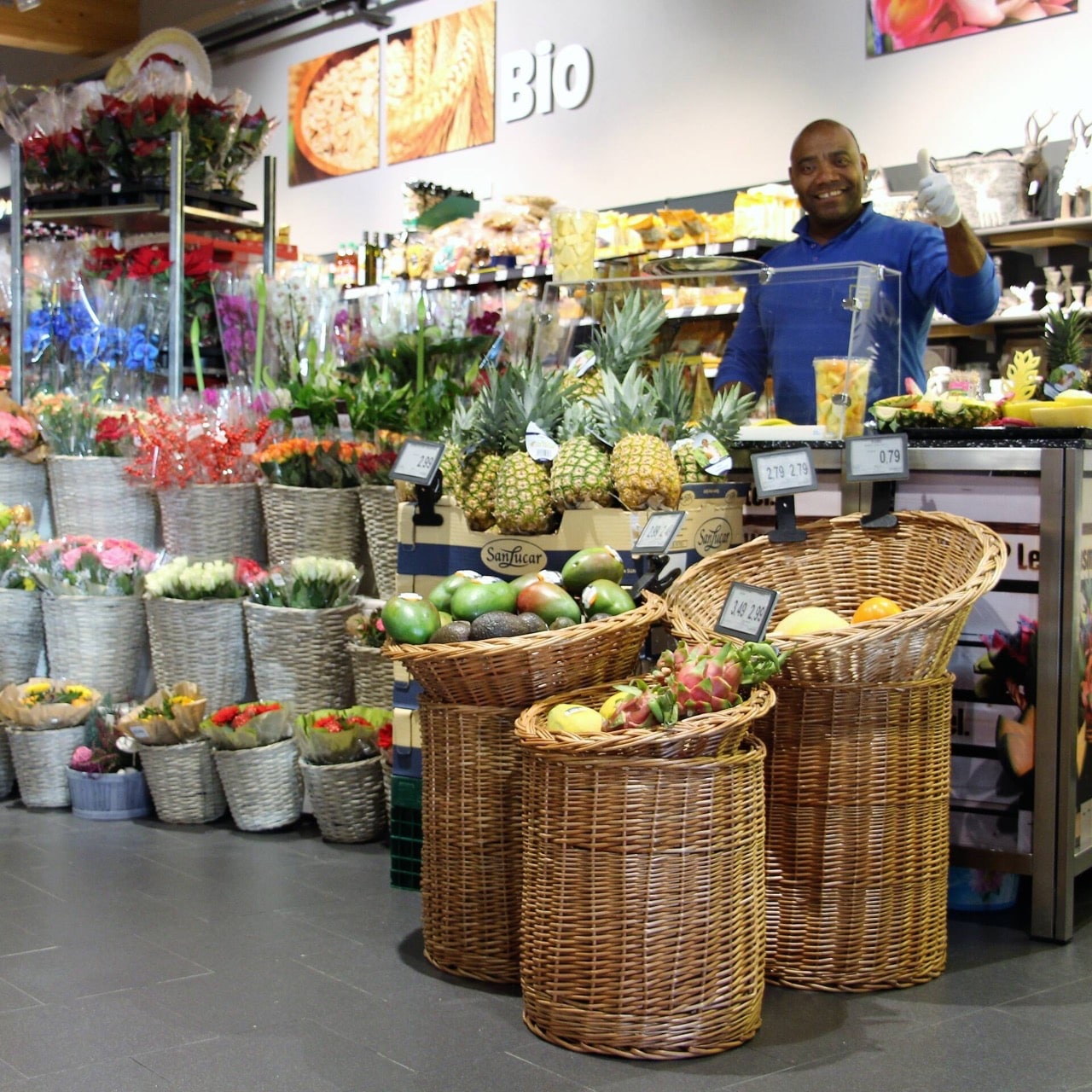 Ein Mann in blauer Kleidung steht hinter einem Tresen. Davor Südfrüchte und Blumen.