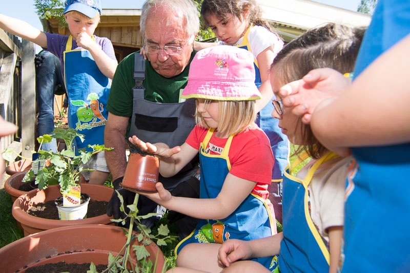 Ein Kind mit Sonnenhut und blauer Schürze schlägt auf den Boden eines kleinen Plastic-Pflanzentopfes. Ein älterer Herr und weitere Kinder stehen dabei.