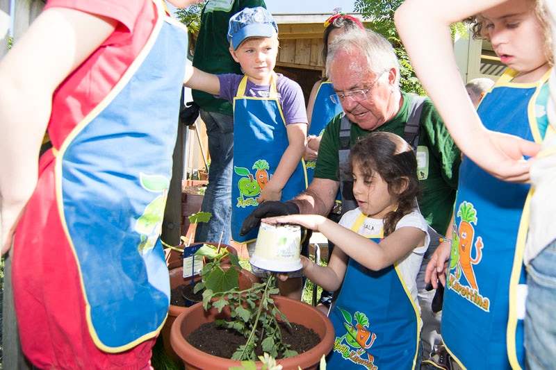Ein Kind mit blauer Schürze schlägt auf den Boden eines kleinen Plastic-Pflanzentopfes. Ein älterer Herr und weitere Kinder stehen dabei.