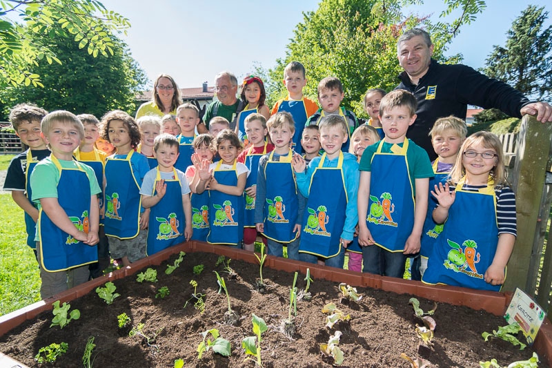 Eine größere Gruppe Kinder mit blauen Schürzen steht an einem Hochbeet. Hinter ihnen stehen drei Erwachsene, einer trägt einen schwarzen EDEKA-Pullover.