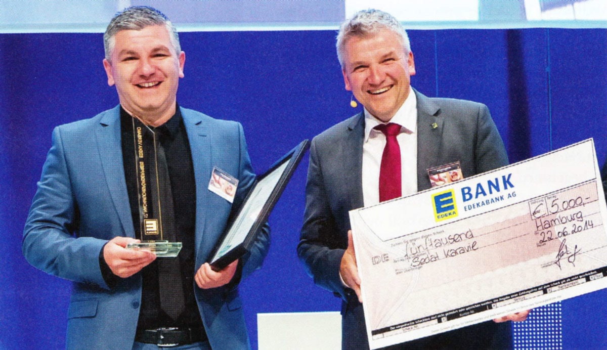 Zwei Männer in Anzügen. Der linke hält eine Auszeichnung in der Hand, der rechte einen überdimensionalen Scheck über 5.000 Euro.