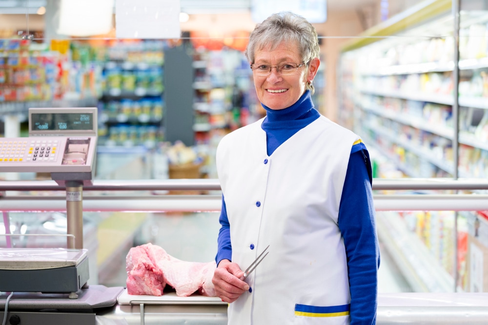 Eine Frau mit grauer Kurzhaarfrisur und Brille hinter der Fleischtheke. Sie trägt einen weißen Kittel und hält eine Fleischgabel in der Hand.