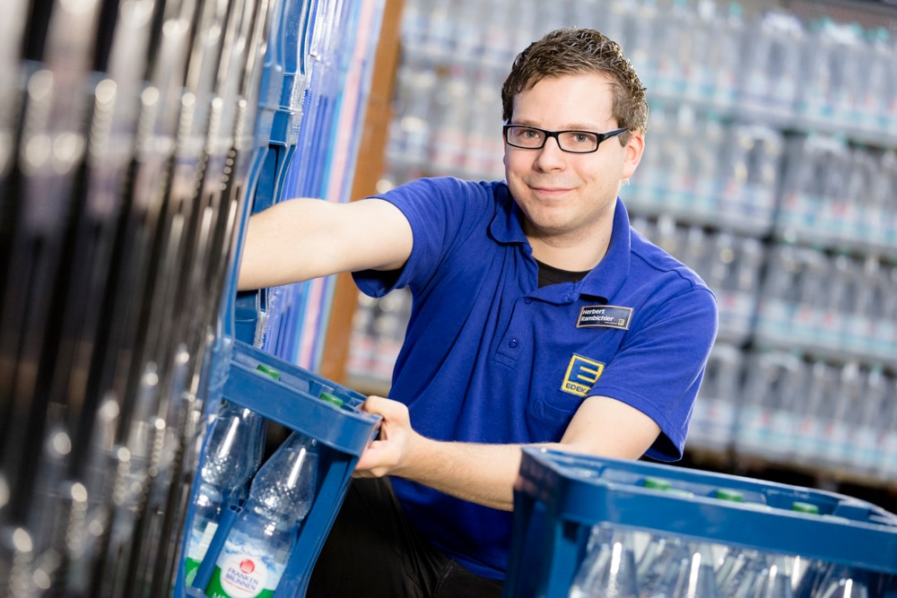 Ein junger Mann mit Brille nimmt einen Wasserkasten aus dem Regal. Er trägt ein blaues EDEKA-Poloshirt.
