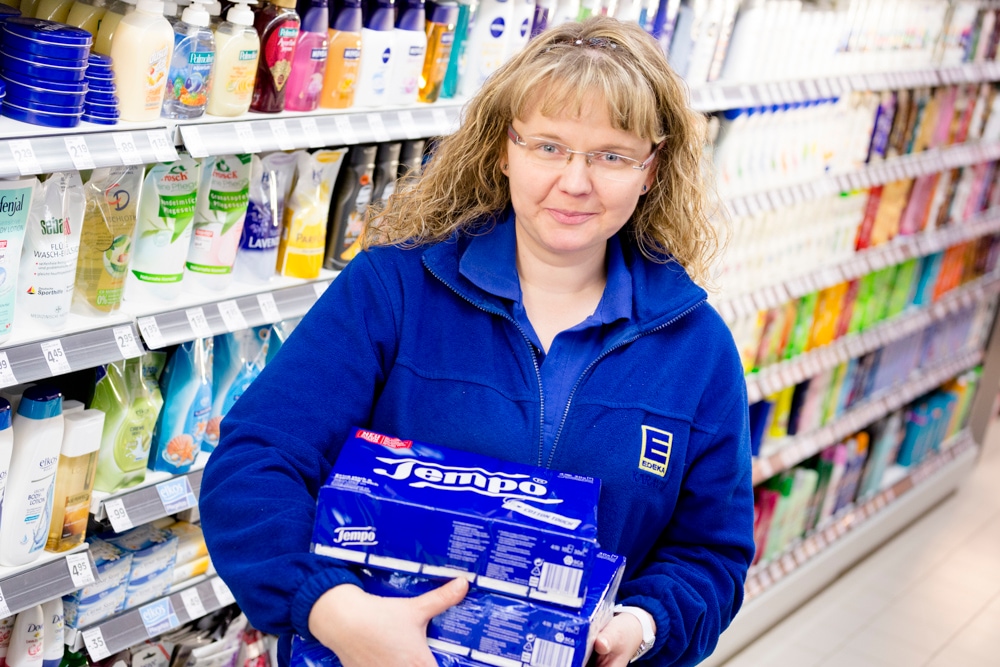 Eine Frau in blauer EDEKA-Kleidung steht in der Drogerieabteilung. Sie hält zwei Großpackungen Tempo-Taschentücher in den Händen.