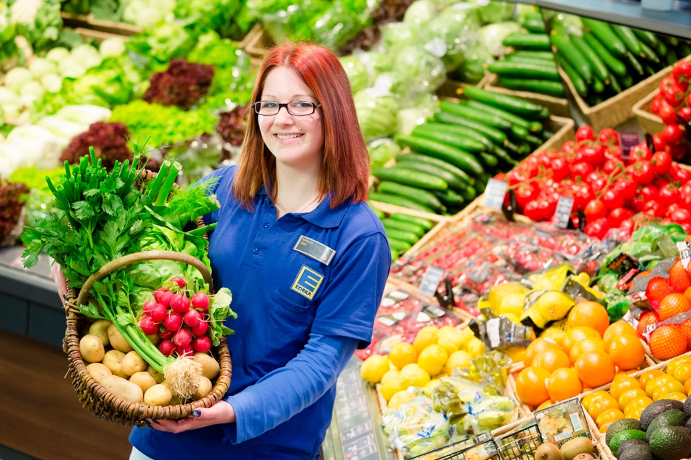 Eine Frau in blauer EDEKA-Kleidung steht in der Obst- und Gemüseabteilung. Sie hält einen Korb mit Kartoffeln, Frühlingszwiebeln und Radieschen in den Händen.