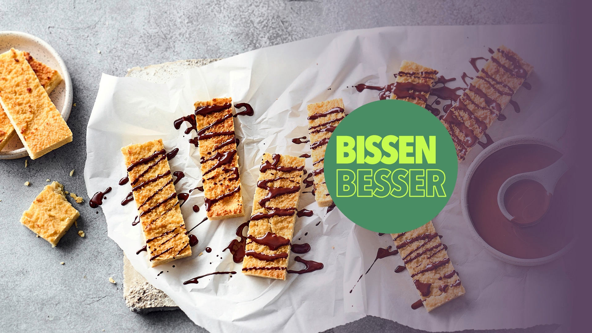 Mehrere Gebäckriegel mit Schokodrizzle liegen auf Backpapier, daneben eine Tasse Kaffee; eingeblendet der Schriftzug „BISSEN BESSER“.