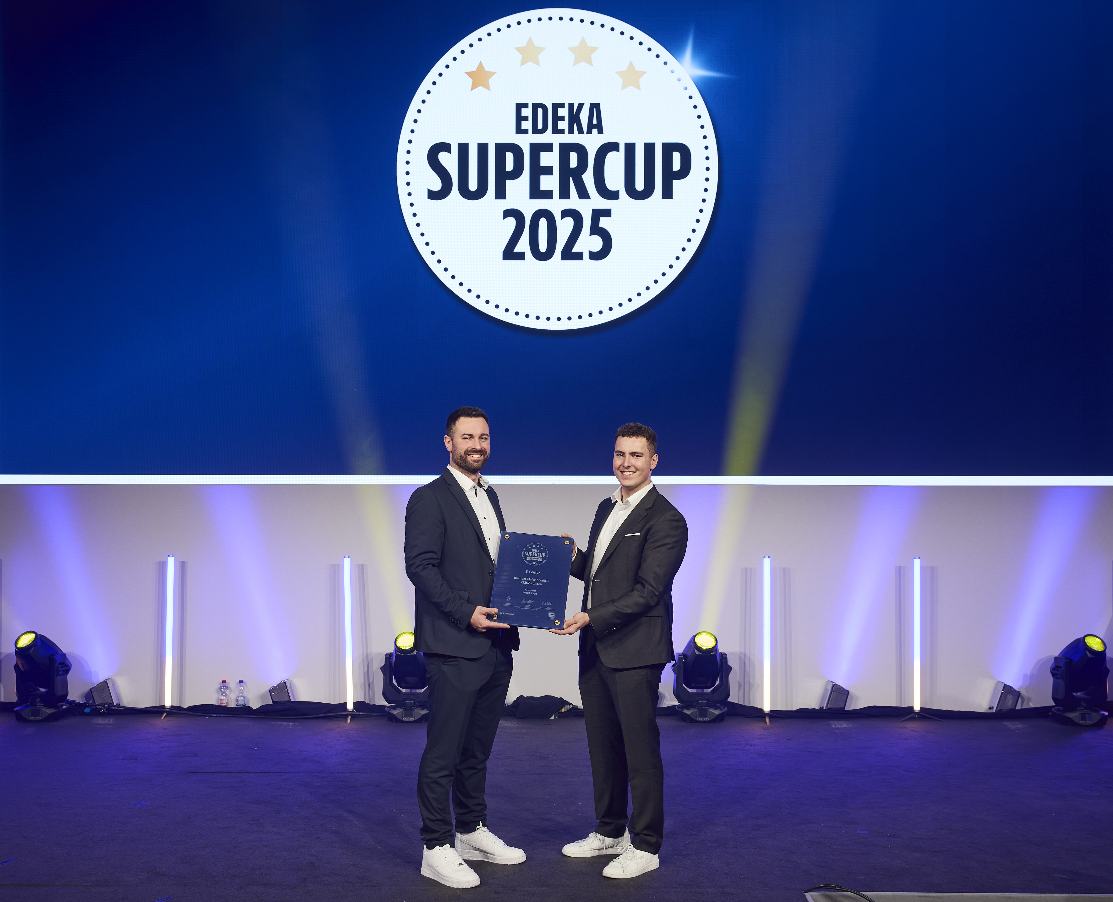 Zwei Männer auf einer Bühne halten eine Urkunde, darüber das Logo „EDEKA Supercup 2025“.