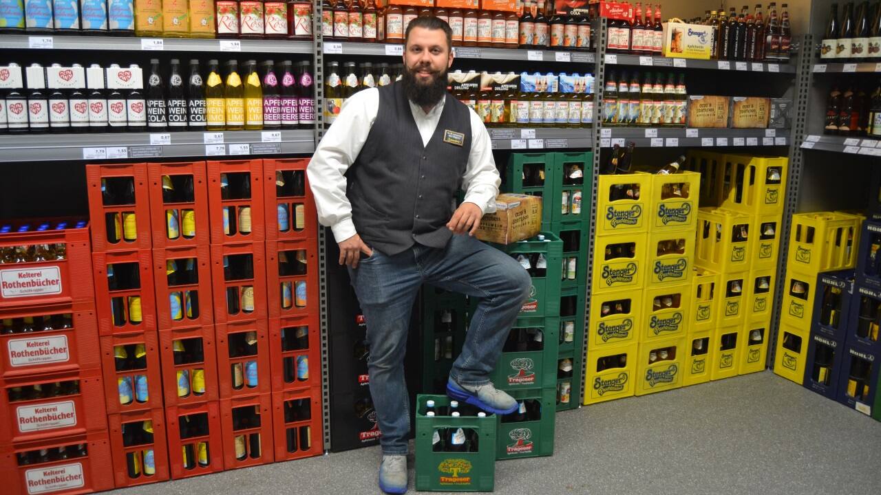 Ein Mitarbeiter eines Getränkemarkts lehnt sich auf eine Kiste und lächelt in die Kamera.
