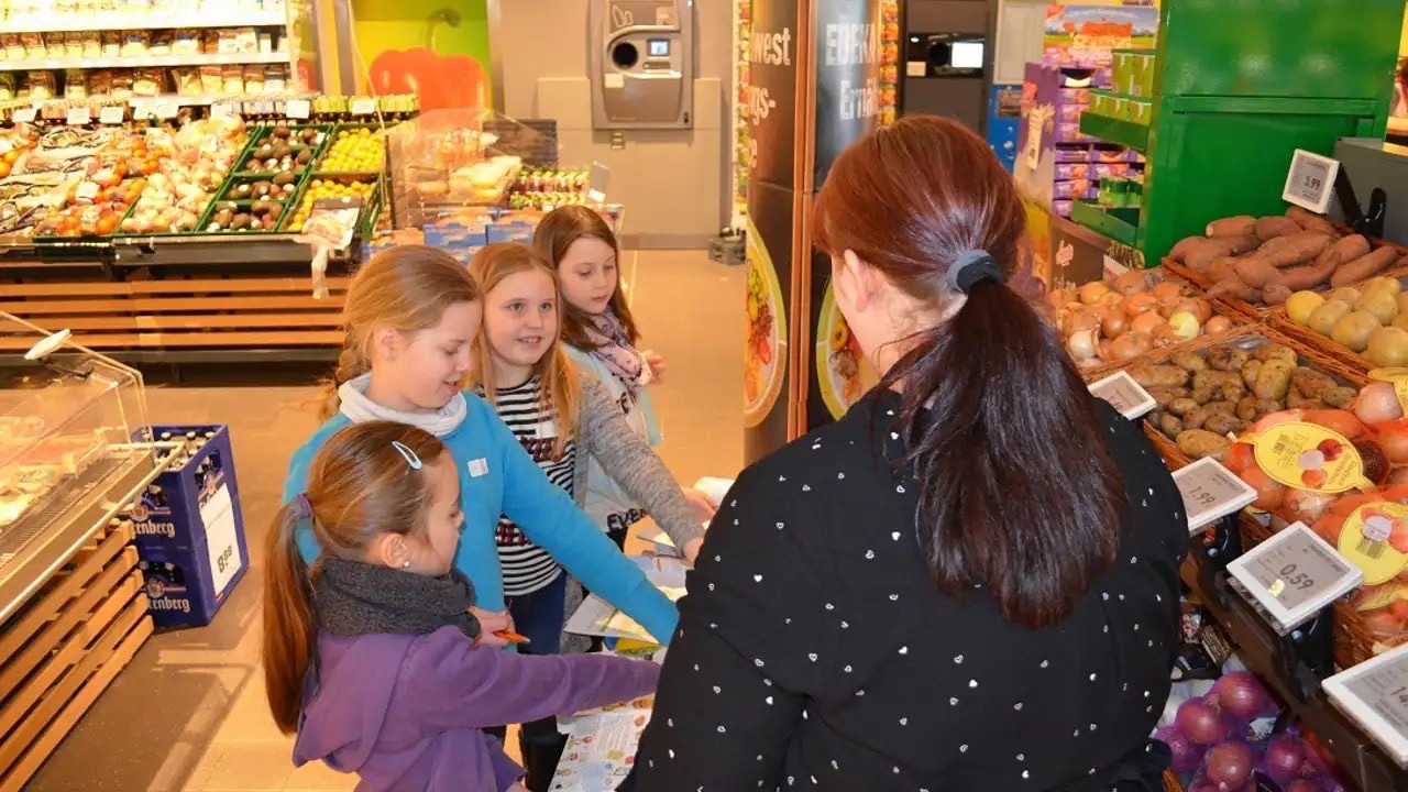 Vier junge Mädchen und eine erwachsene Frau stehen am Gemüseregal eines Einkaufsmarktes.