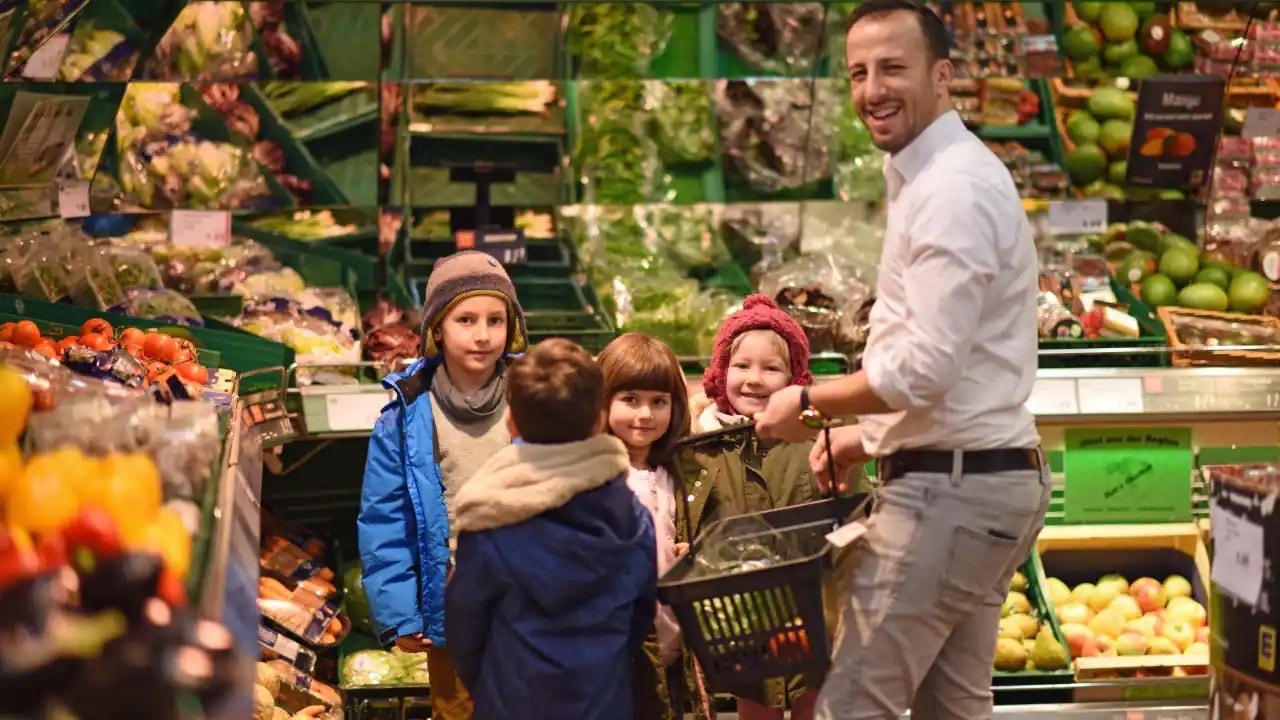 Ein Mann in weißem Hemd mit einem Einkaufskorb steht in der Gemüseabteilung eines Einkaufsmarktes. Um ihn herum vier kleine Kinder.