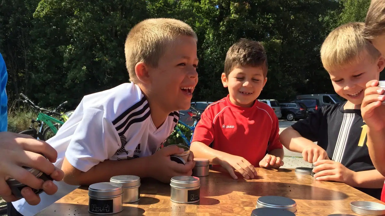Drei kleine lachende Jungs draußen an einem Tisch auf dem Metalldosen stehen.