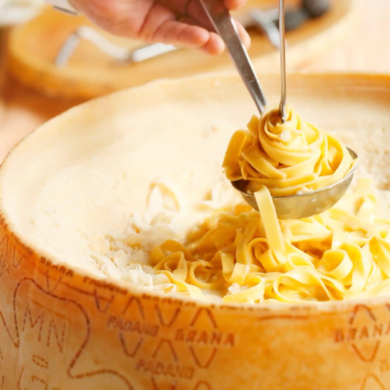 Bandnudeln werden mit einer Gabel in einer Metallkelle aufgerollt. Unter der Kelle Bandnudeln in einem ausgehöhlten Käselaib.