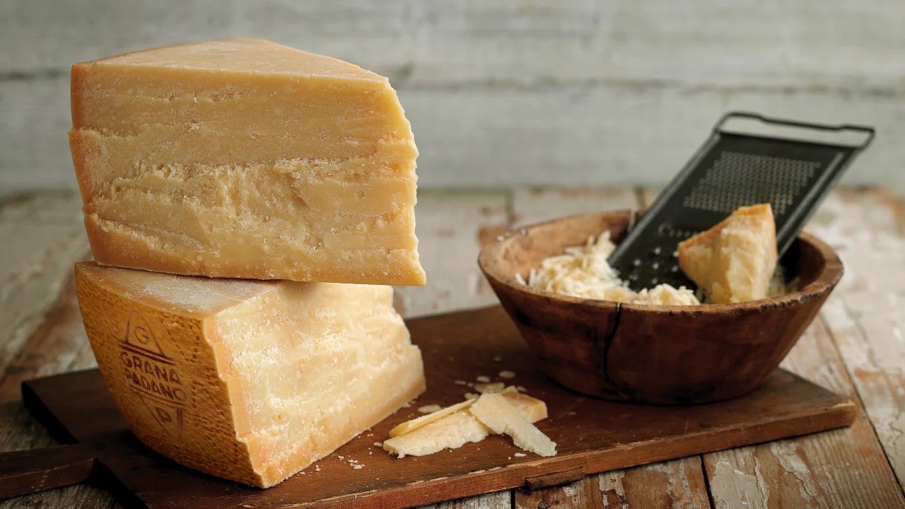 Zwei große Dreiecke Parmesan Hartkäse auf einem braunen Holzbrett. Daneben ein Schälchen mit einer Handreibe.