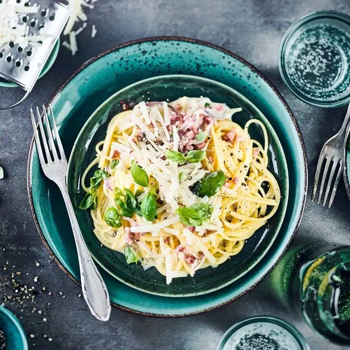 Ein Teller Spaghetti mit weißer Soße, geraspeltem Parmesan und Basilikumblättern.