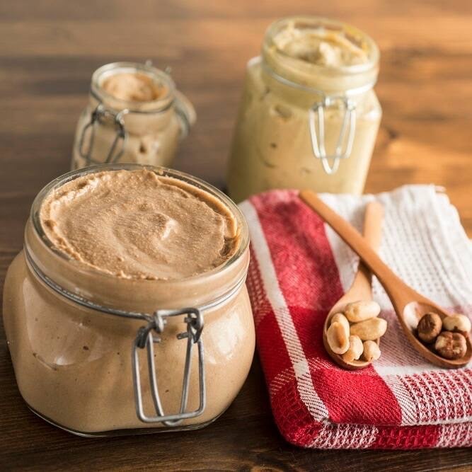 Einmachglas mit einer hellbraunen Masse. Daneben zwei kleine Holzlöffel mit Nüssen auf einem Geschirrtuch.