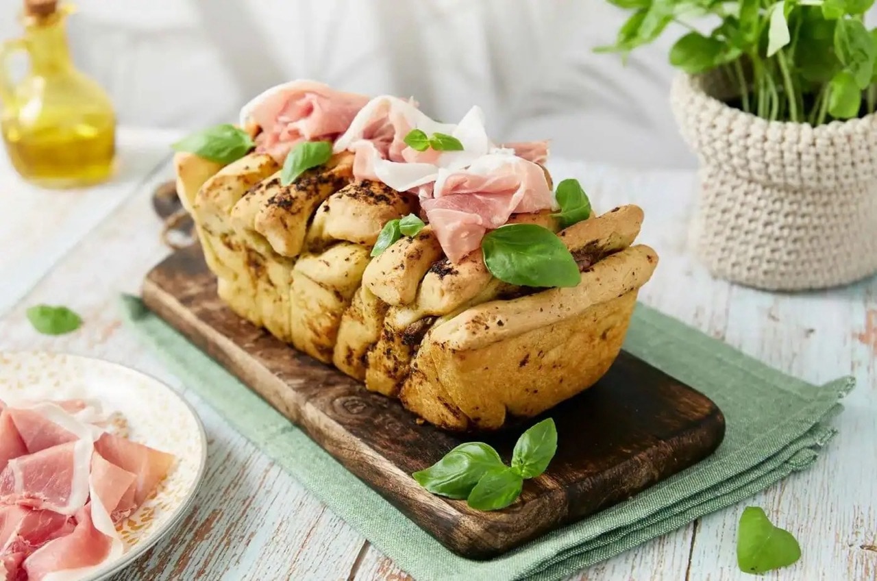 Längliches Fladenbrot auf einem Holzbrett. Auf dem Brot liegen Schinkenscheiben und grüne Basilikumblätter.