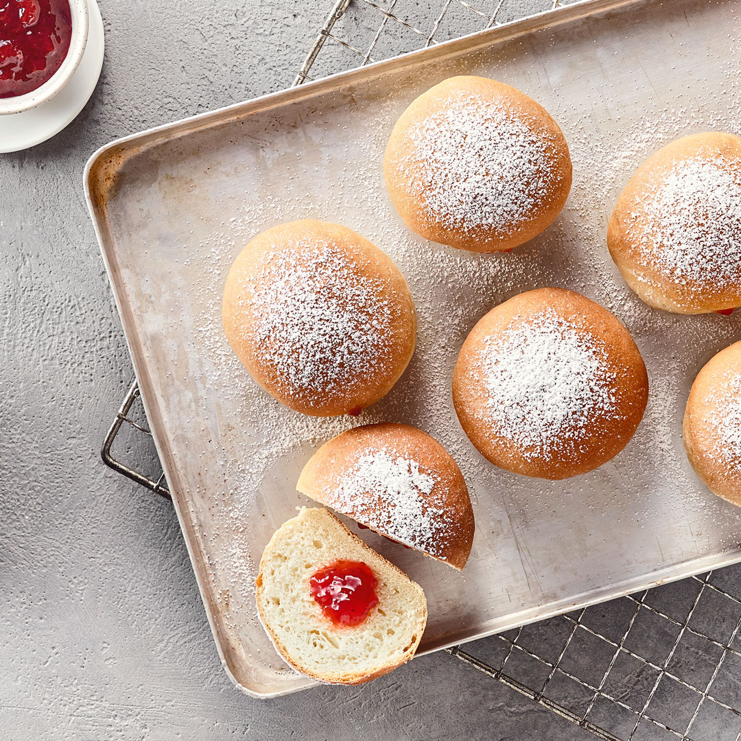 Ein Backblech mit gebackenen Teigstückchen, davon eins aufgeschnitten & mit roter Marmelade gefüllt, garniert mit Puderzucker