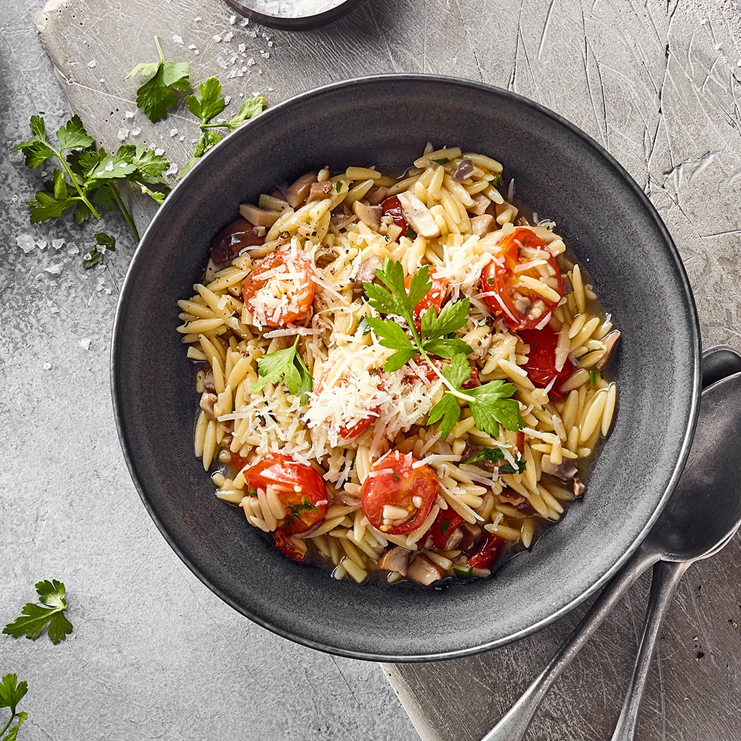 Eine Schale mit Orzonudeln, Kirschtomaten und Champignons, getoppt mit geriebenem Käse und frischer Petersilie