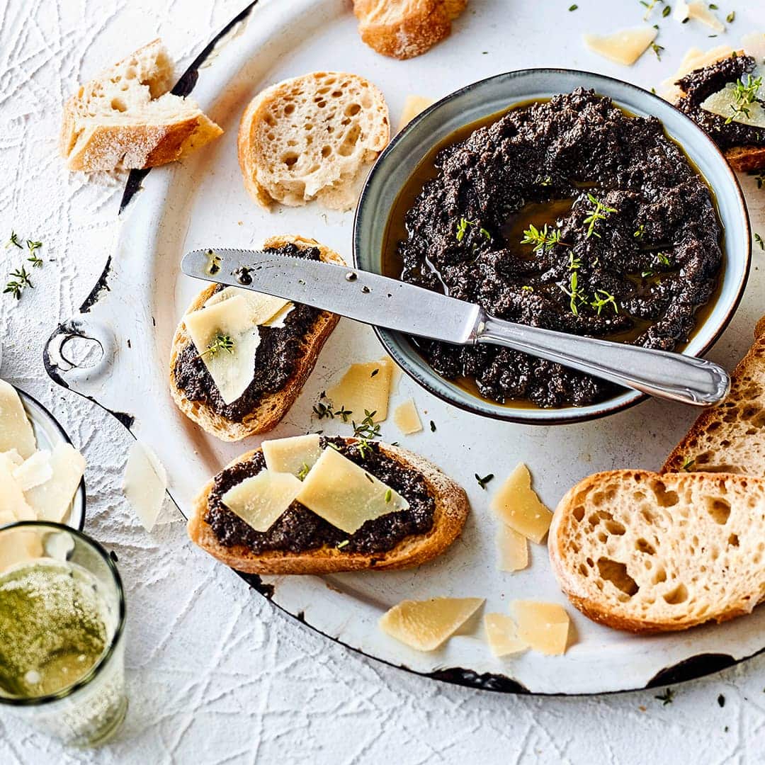 Eine Paste aus schwarzen Oliven, Knoblauch, Sardellen und Olivenöl ist ein würziger Brotaufstrich für Baguette. Garnieren Sie das Ganze noch mit Parmesan und Thymianblättchen!