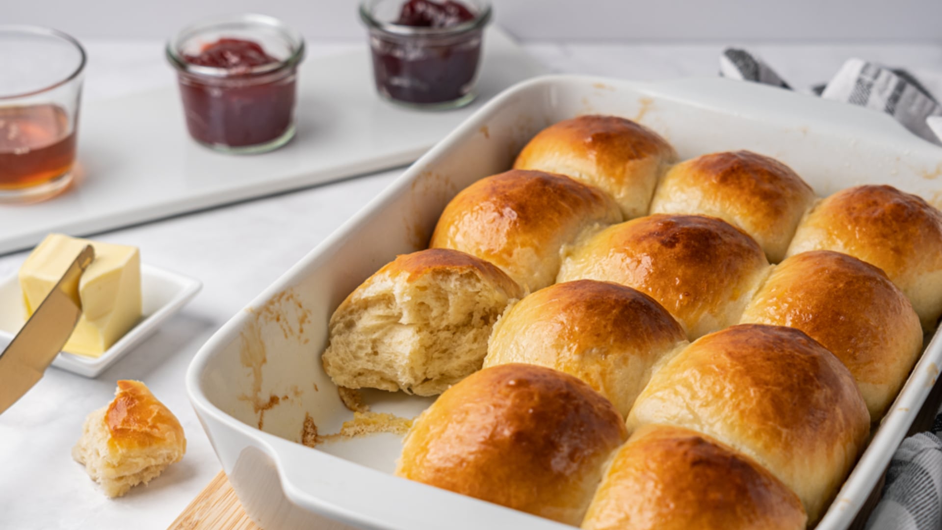 Eine Auflaufform mit goldbraun gebackenen Brötchen, daneben Schüsseln mit Konfitüre und Butter