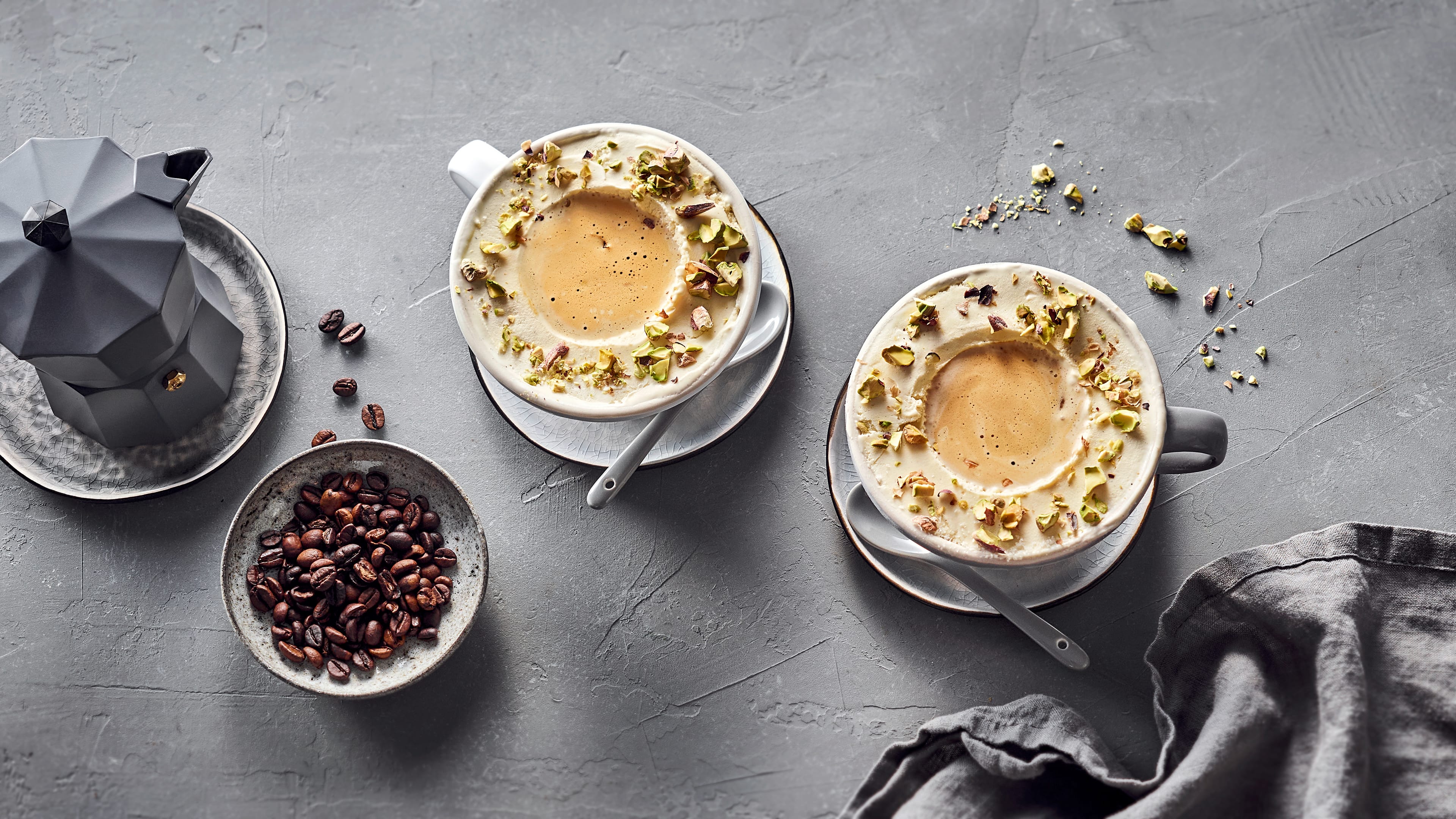Runde große Kaffeetassen mit luftigem, hellem Eis, in der Mitte cremiger Kaffee, getoppt mit gehackten Pistazien