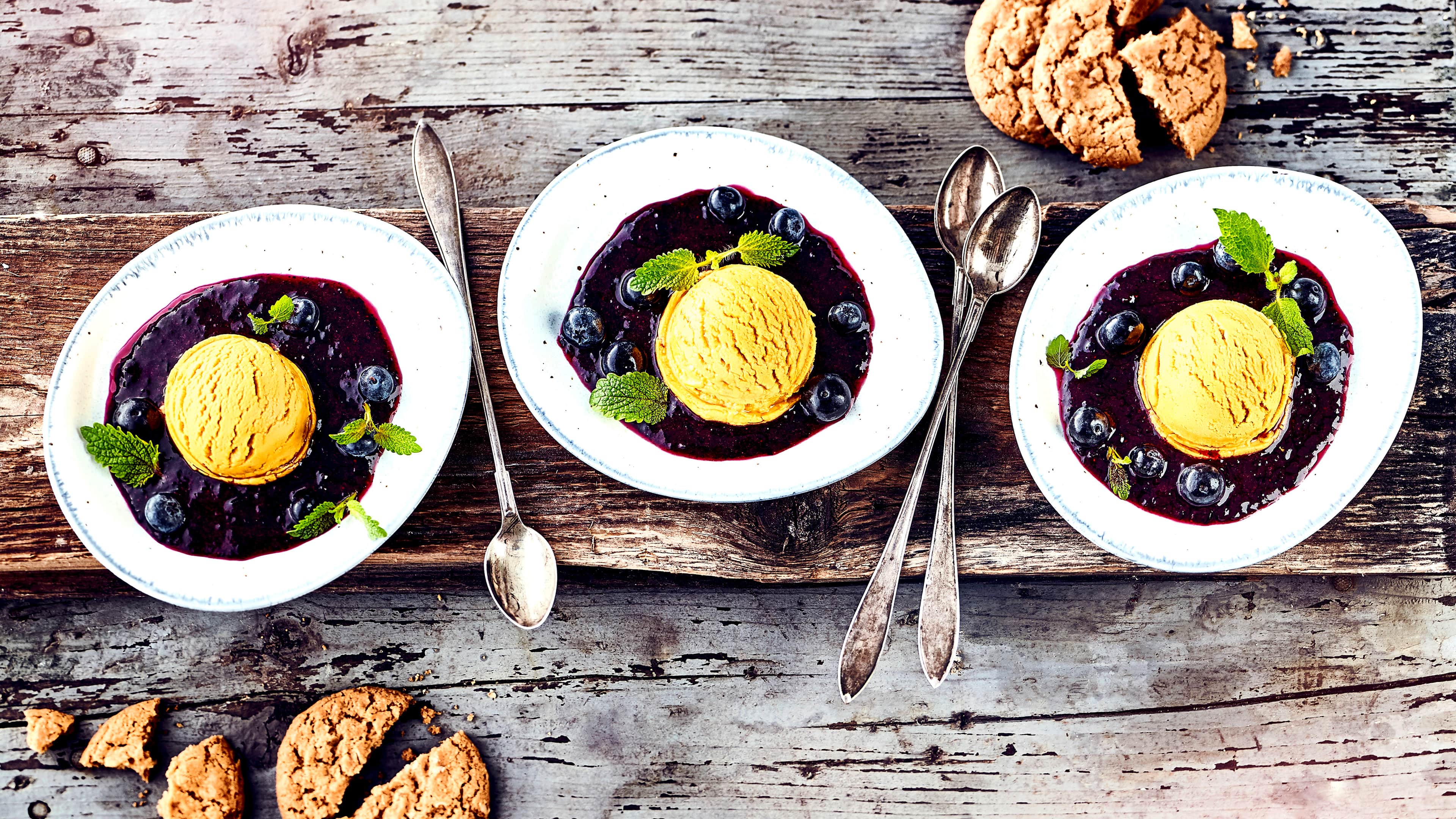 Rezept für Aprikosensorbet mit Soße Ovale Schalen mit gelborangenem Sorbet, in einer dunklen Beerensoße mit Blaubeeren, getoppt mit frischen Melisseblättchen