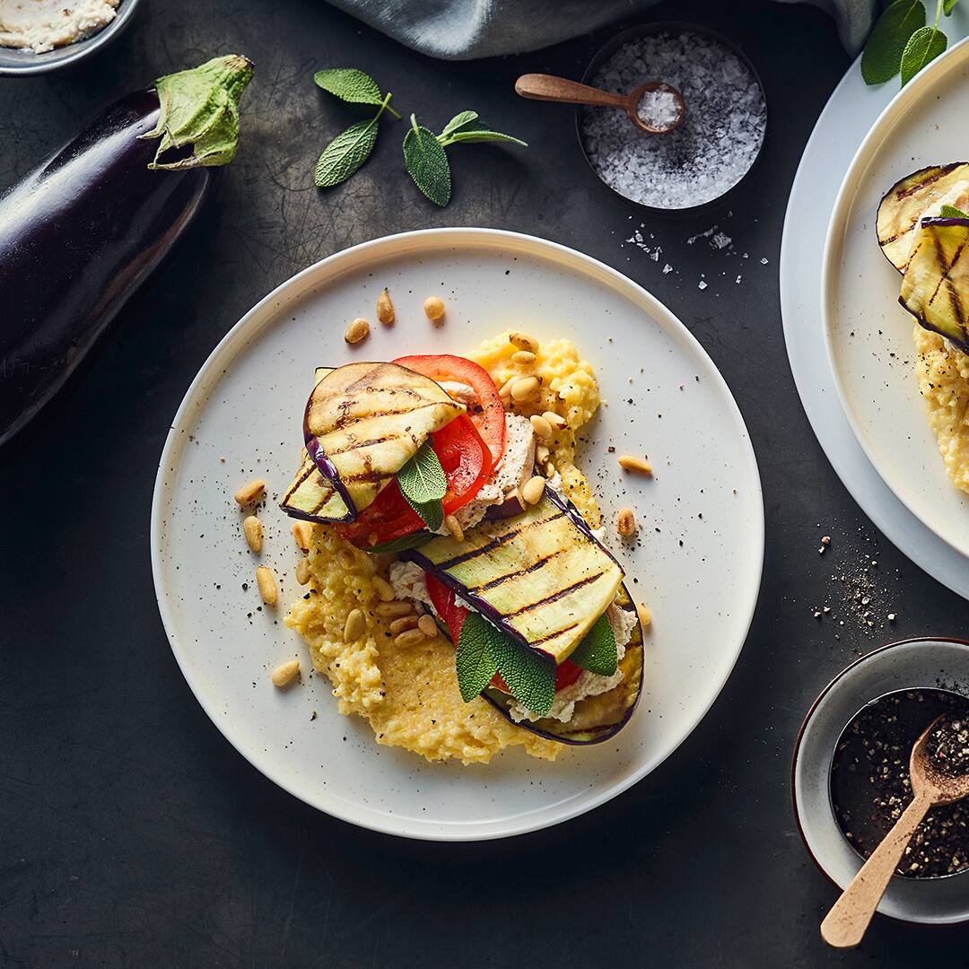 Gegrillte Auberginenscheiben mit Ricottacreme und Tomatenscheiben auf einem Bett aus Polenta, garniert mit Pinienkernen