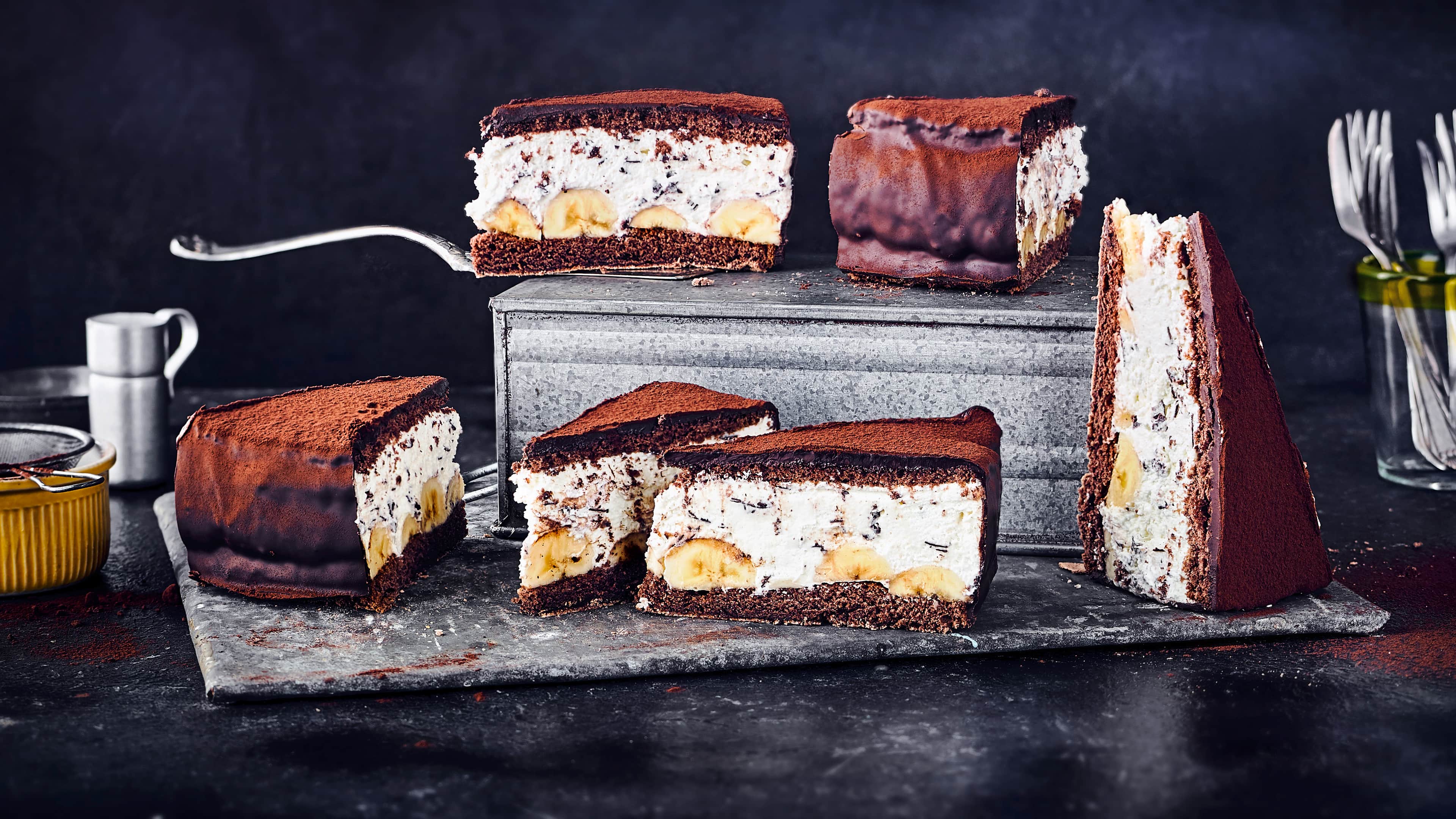 Rezept für eine Schoko-Bananentorte Ein Metallblech mit Tortenstücken mit Schokoböden, Straciatella-Creme und Bananenstücken, bestreut mit Kakao