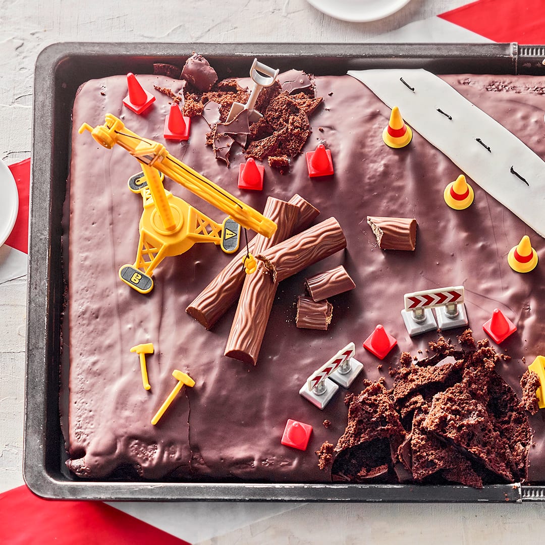 Ein Schoko-Blechkuchen mit Zartbitter-Kuvertüre, der an zwei Stellen aufgebrochen, dekoriert mit Spielzeug-Bagger und Hütchen