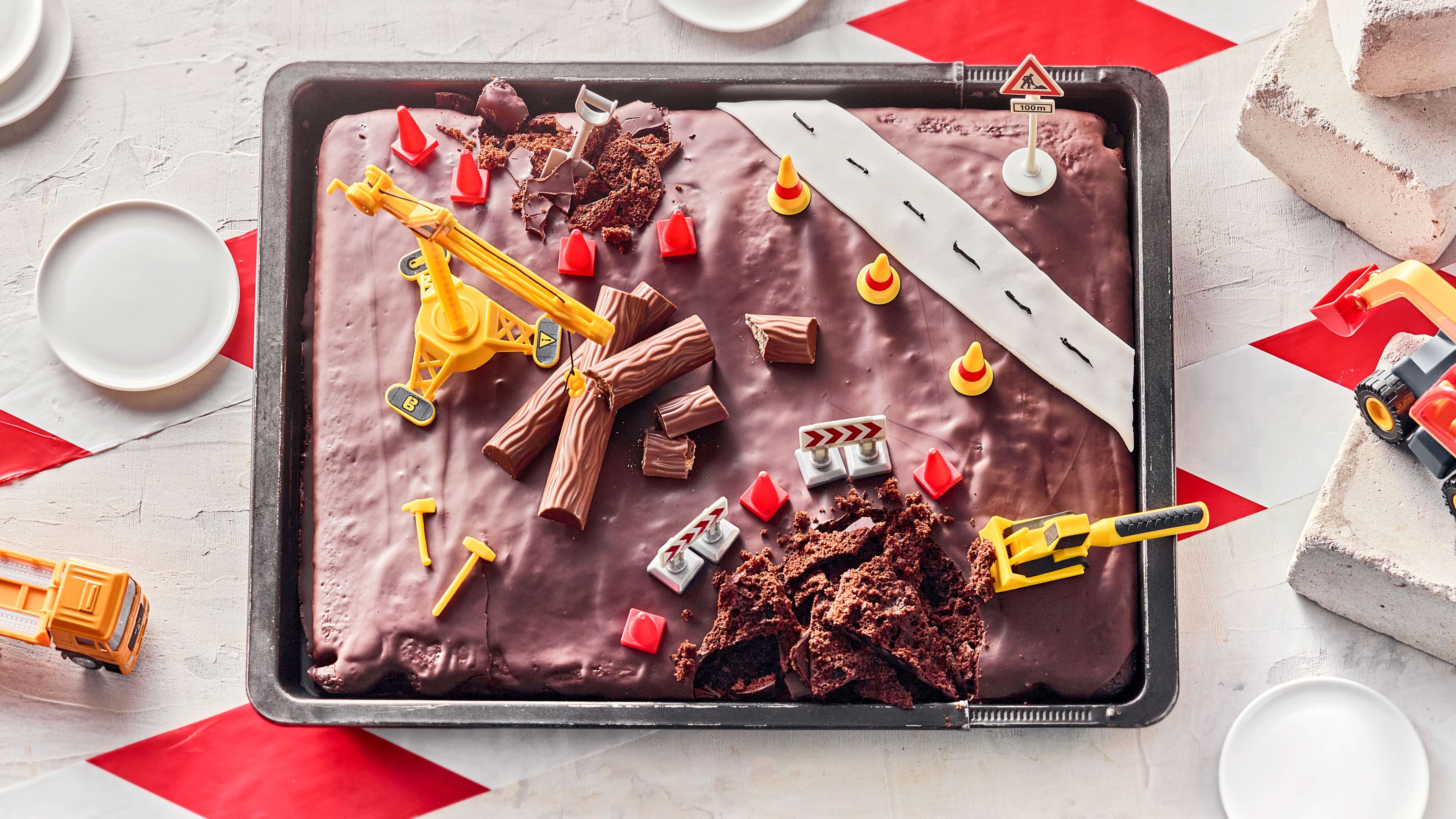 Rezept für Baustellen-Kuchen Ein Schoko-Blechkuchen mit Zartbitter-Kuvertüre, der an zwei Stellen aufgebrochen, dekoriert mit Spielzeug-Bagger und Hütchen