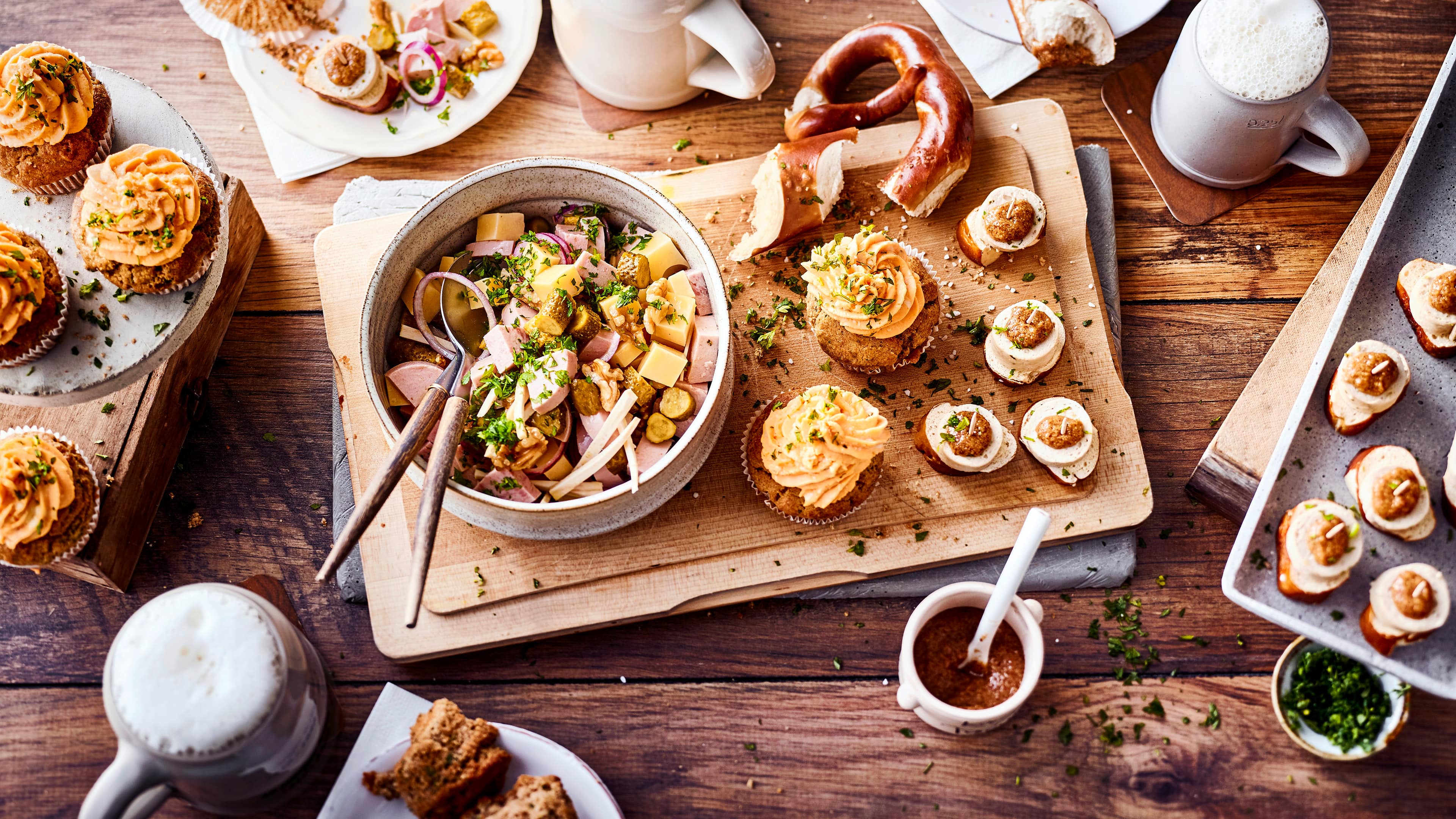 Rezept für bayerische Tapas Ein Holzbrett mit herzhaften Cupcakes mit Käse-Creme, Weißwurst-Laugen-Häppchen und eine Schale mit Käse-Wurst-Salat