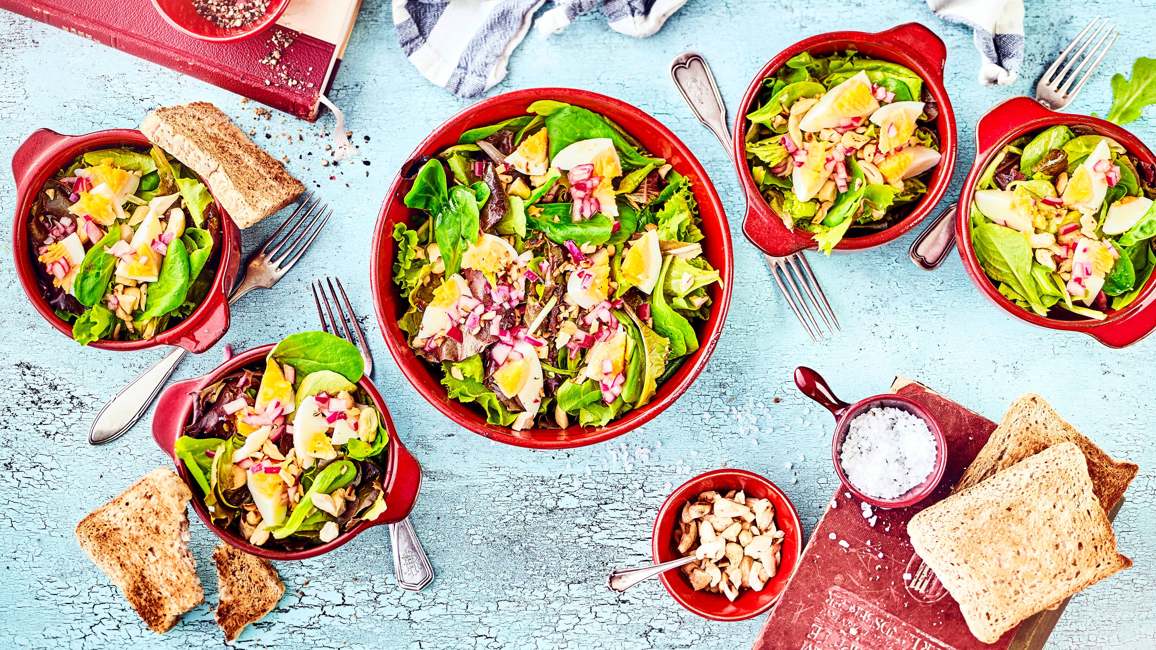 Rezept für Blattsalat mit Cashewkernen Schüsseln mit Blattsalat, gekochten Eiern, roten Zwiebeln und gerösteten Cashewkernen, dazu Vollkorntoast
