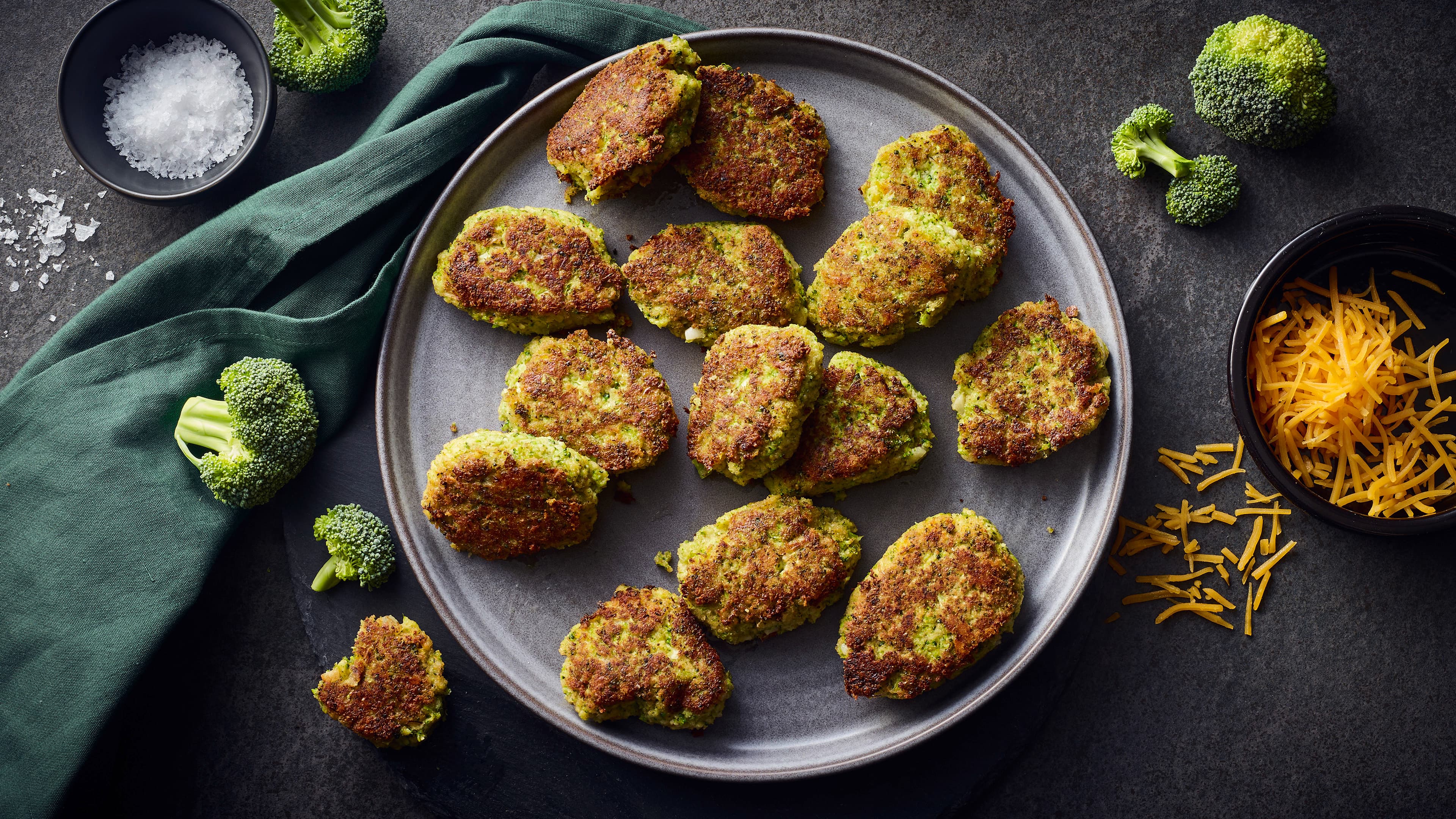 Rezept für knusprige Brokkoli Nuggets Ein dunkler Teller, darauf kleine knusprig gebratene grüne Nuggets, daneben Brokkoli Röschen und eine Schale mit grobem Salz