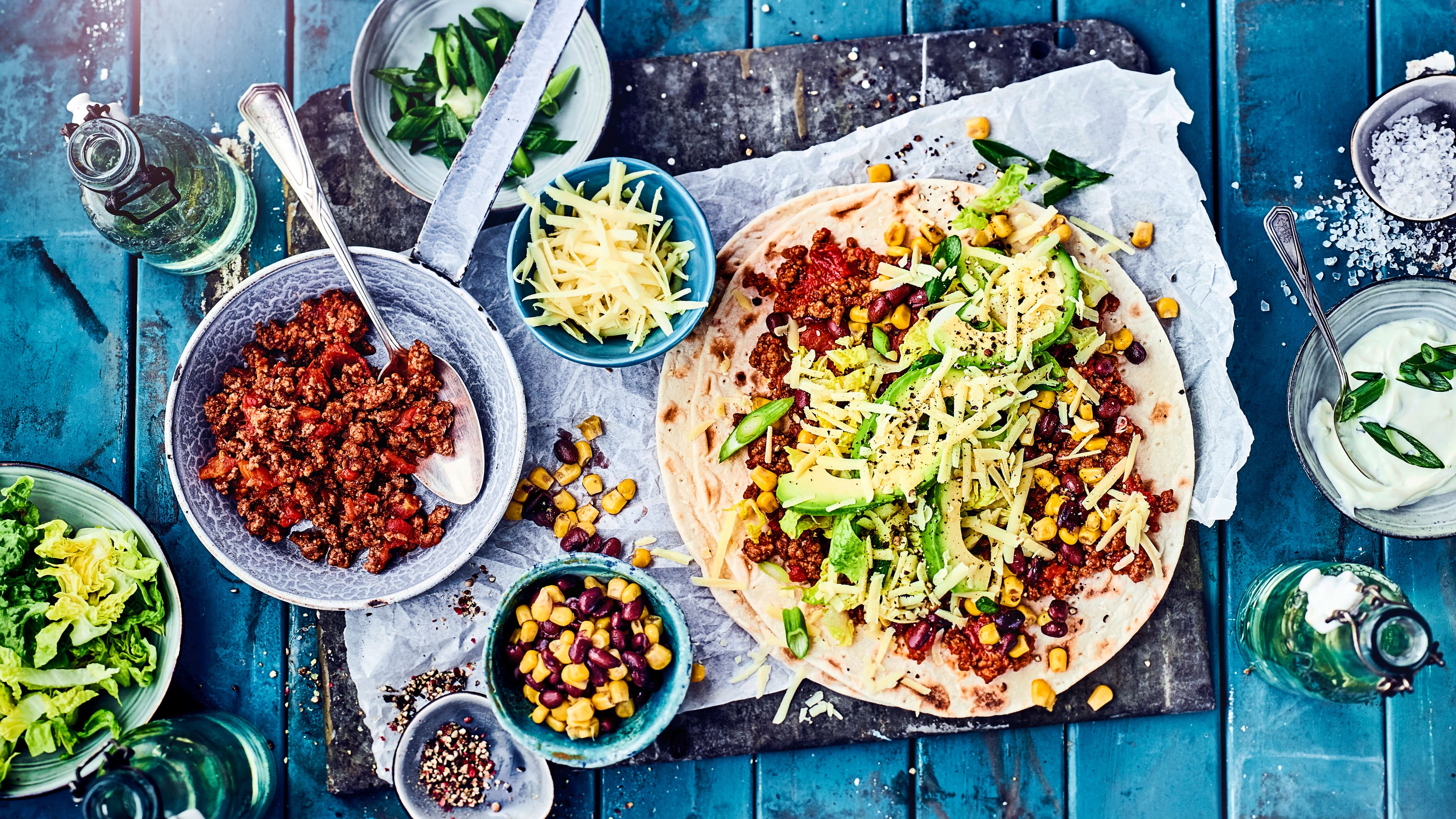 Rezept für mexikanische Burrito Eine Steinplatte mit einem Wrap, gefüllt mit Hackfleisch, Mais, Kidneybohnen, Avocado, Salat und geriebenem Käse