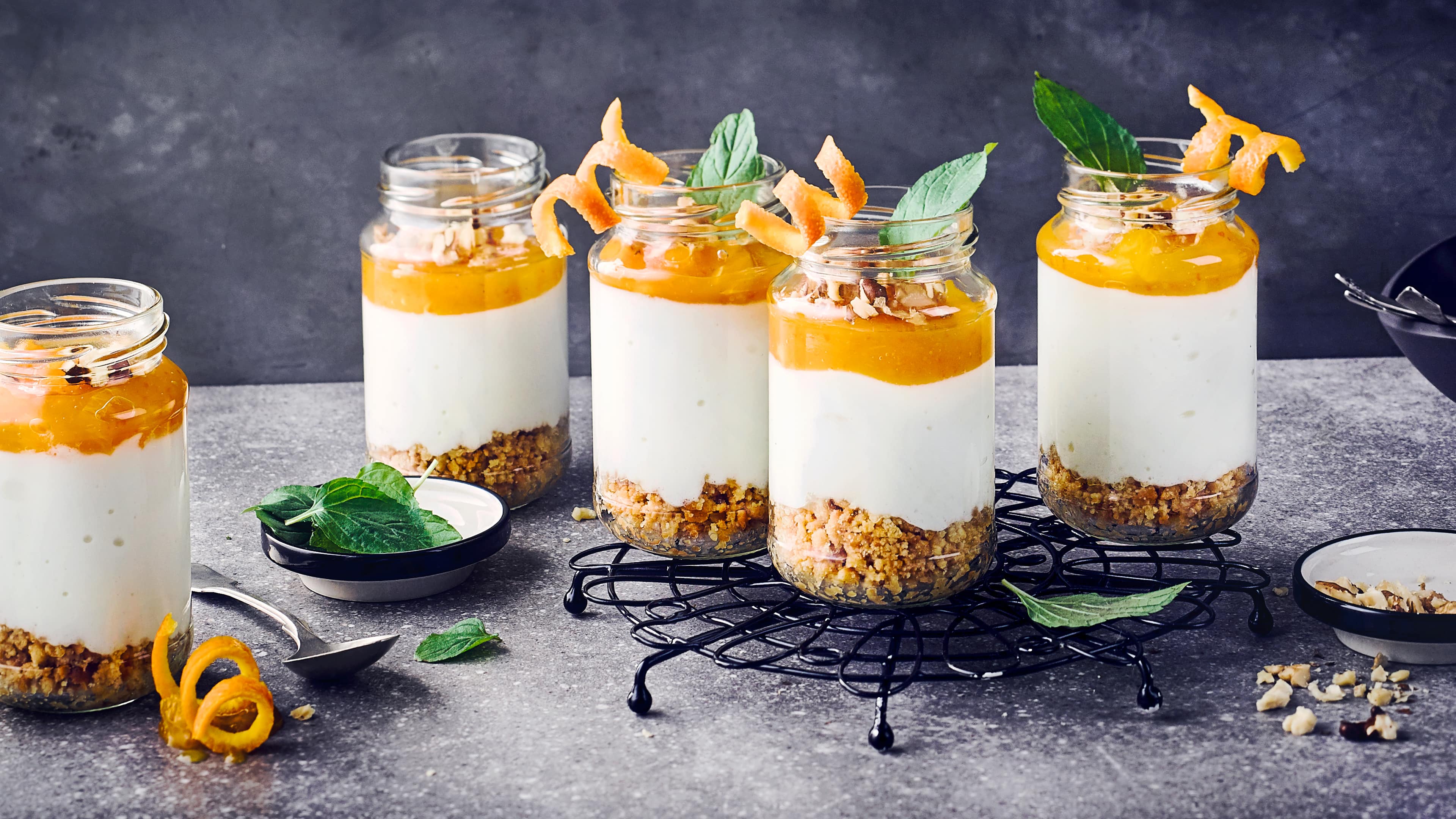 Rezept für Cheesecake im Glas Mehrer Einmachgläser darin geschichtet sind Krümel, eine weiße und eine orangene Creme, getoppt mit Minze und Orangenzeste