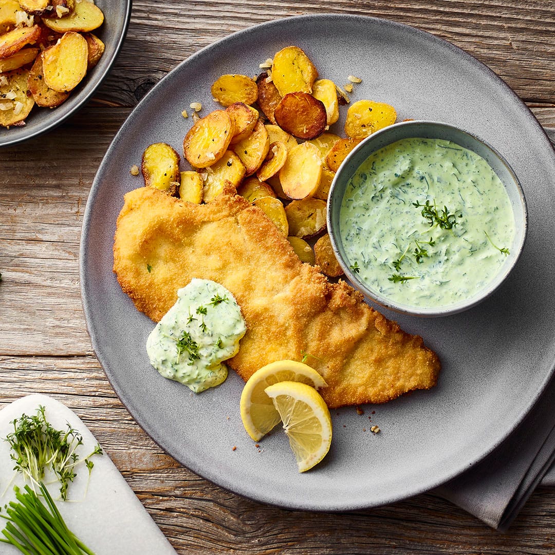 Ein Teller mit frittiertem Schnitzel, Bratkartoffeln, grüne Soße in einem Schälchen, getoppt mit Petersilie und Schnittlauch